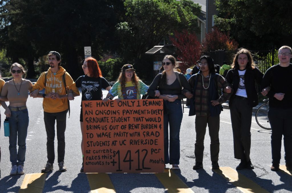 Students linked arms across a crosswalk. One has a large sign that reads: "We have only 1 demand: An ongoing payment to every graduate student that would bring us out of rent burden and to wage parity with grad students at UC Riverside. At current rates this amounts to +$1412 per month."