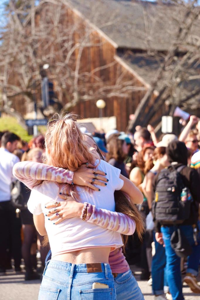 Two protesting students hug in the intersection, with a crowd of people and the Barn Theater in the background