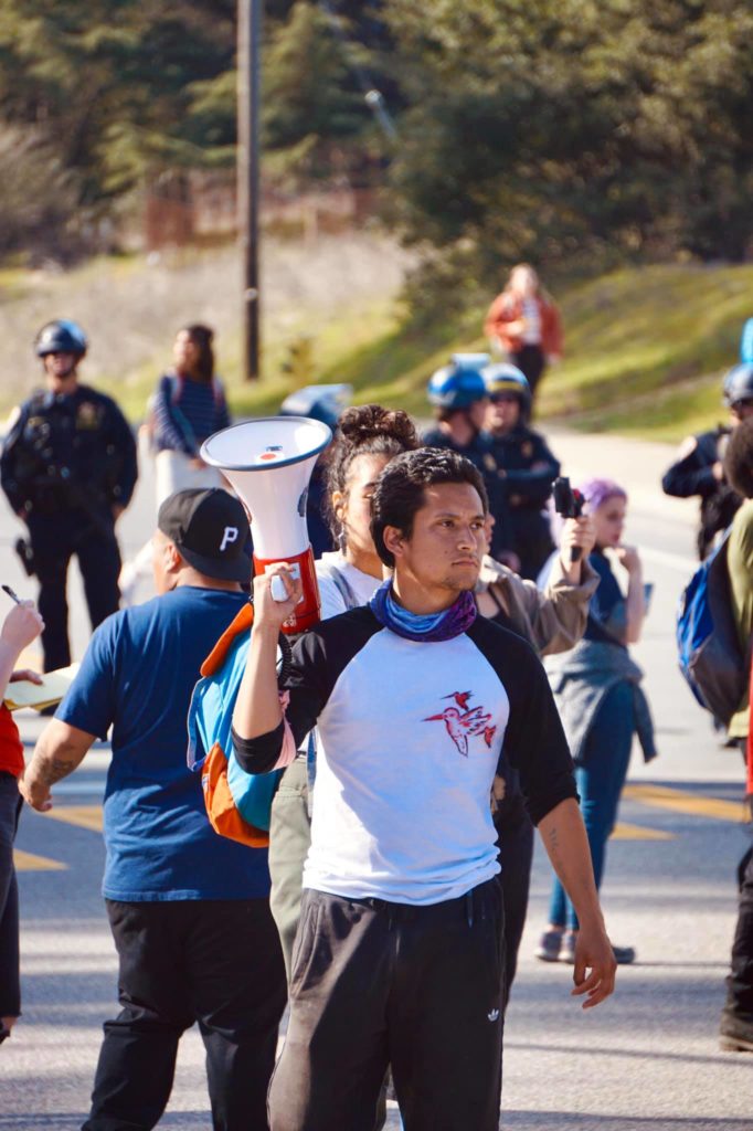 A student with a megaphone standing in the intersection with cops in the background