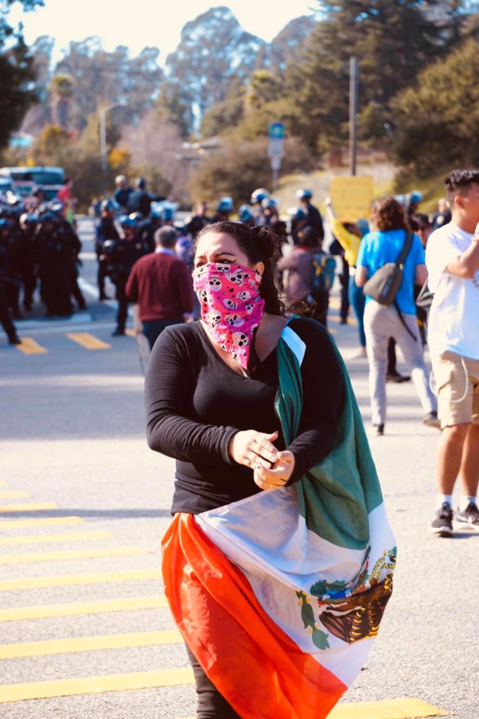 A protester draped in a Mexican flag walks in the intersection, with a large group of police in the background