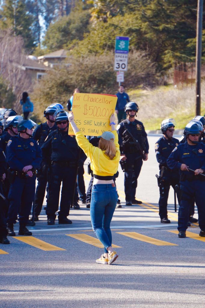 A student holding a sign up to a large group of police. The back of the sign reads "Our Chancellor earns $500,000 / Does she work harder than you?"